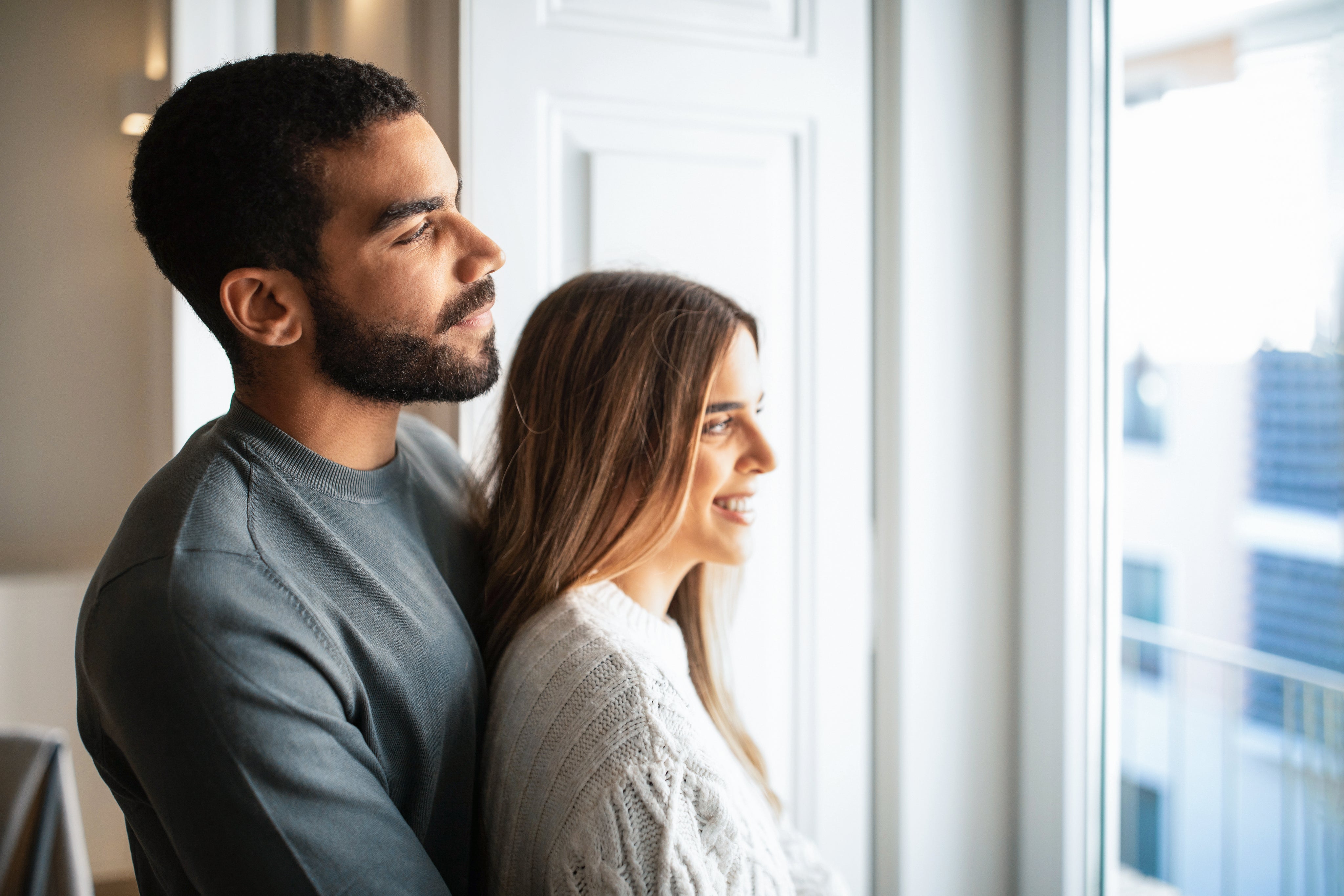 Man and woman standing close together by a window, looking out.